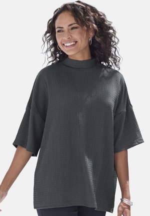 Smiling woman with curly hair wears a textured dark oversized short-sleeve top and silver hoop earrings and bracelets against a plain background.