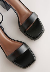 Czarne skórzane sandały na obcasie z szerokim paskiem i kwadratowym noskiem. Brązowa wkładka z kontrastującymi przeszyciami. Obcas typu blok.