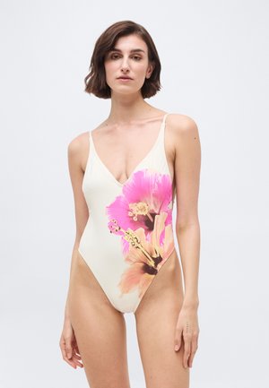 Sieviete ar īsiem brūniem matiem, tērpusies baltā viengabala peldkostīmā ar lielu rozā un persiku ziedu rakstu priekšpusē.