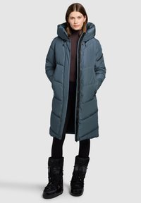 Gepolsterter, langer Mantel in Schieferblau mit Kapuze, Reißverschluss und Seitentaschen. Kombiniert mit schwarzen, wasserdichten Stiefeln bis zur Mitte der Wade.