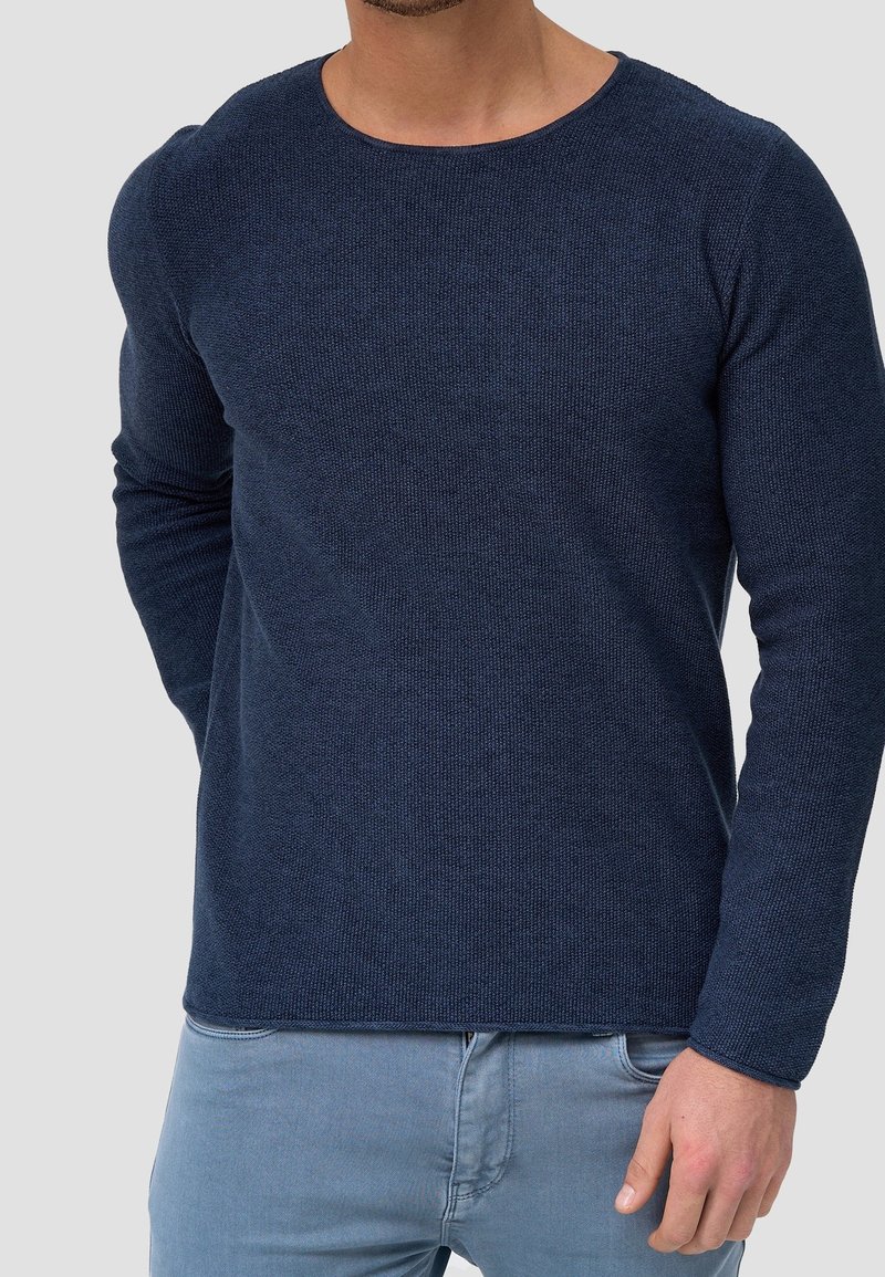 Homme portant un pull à manches longues bleu marine texturé et un jean bleu clair, debout contre un fond uni gris clair.