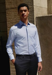 Helles blaues langärmliges Hemd mit Button-Down-Kragen, einfacher Brusttasche, kombiniert mit dunklen strukturierten Anzughosen.