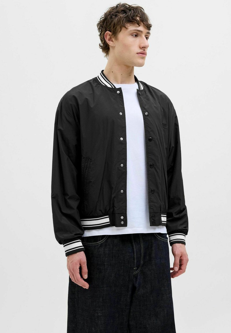 Blouson noir avec col et poignets en ribbed à rayures blanches, doté d'une fermeture à boutons-pression, de poches latérales et d'une texture lisse.