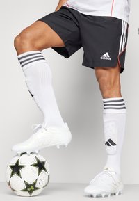 Baltie futbola apavi ar melniem akcentiem, melni šorti ar sarkaniem ieliekumiem un baltas ceļgala augstuma zeķes ar melnām svītrām, atpūšas uz zvaigžņu rakstu futbola bumbas.
