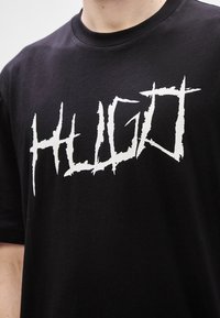Μαύρη βαμβακερή μπλούζα με κοντό μανίκι που διαθέτει ένα μεγάλο, φθαρμένο άσπρο λογότυπο "HUGO". Σχέδιο με στρογγυλή λαιμόκοψη και κοντά μανίκια. Η υφή φαίνεται μαλακή και φιλόξενη.