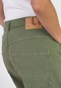 Pantalones cargo verde oliva con un parche de cuero en la parte trasera, dos bolsillos traseros y costuras decorativas a lo largo de las costuras y bordes de los bolsillos.