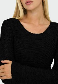Pull en maille noir avec un col rond large et des manches longues, présentant une finition texturée. Le tissu semble doux et épais.