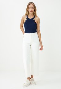 Jonge vrouw met lang blond haar, gekleed in een marineblauwe mouwloze ribbed top met gouden knopen, witte wijde broek en witte sneakers.