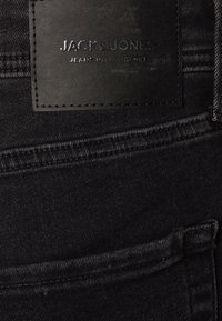 Černé denimové džíny s texturovaným povrchem, s černou koženou nášivkou vyraženou s nápisem "JACK & JONES" a viditelnými detaily šití.