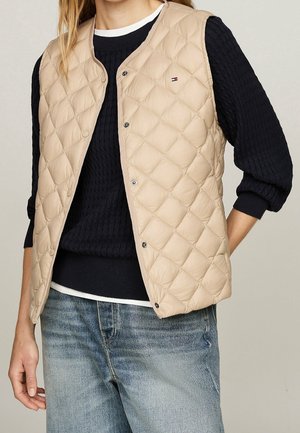 Nainen, joka pukeutunut beigenväriseen tikattuun liiviin, jossa on painonapit, mustan kaapelineulospuseron ja kuluneiden sinisten farkkujen päälle.