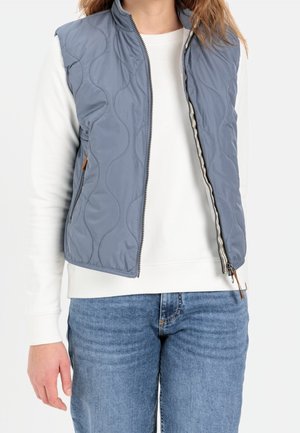 Personne portant une veste matelassée bleu clair sur une chemise blanche à manches longues et un jean bleu, veste ouverte à mi-hauteur.