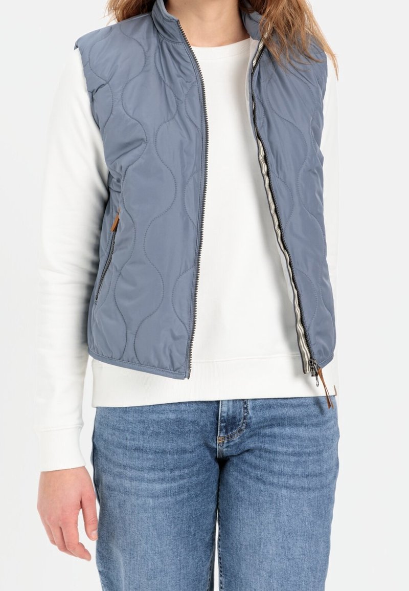 Personne portant une veste matelassée bleu clair sur une chemise blanche à manches longues et un jean bleu, veste ouverte à mi-hauteur.