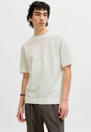 Ung mand med mørkt, bølget hår iført en tekstureret hvid kortærmet sweater og mørkegrå bukser mod en ensfarvet baggrund.