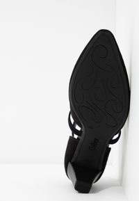 Chaussure à talon haut en cuir de daim noir avec un bout pointu et un design strappy décoratif, présentant une semelle en caoutchouc texturé et un logo embossé.