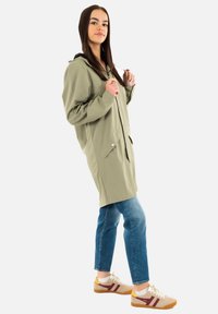 Impermeable verde oliva claro con capucha, botones a presión y bolsillos frontales. Combinado con jeans de mezclilla azul y zapatillas deportivas beige con detalles en burdeos.