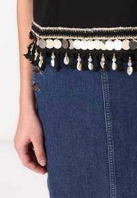 Top negro con acentos circulares metálicos y borlas de cuentas, combinado con una falda de mezclilla azul de tiro alto con detalles de botones.