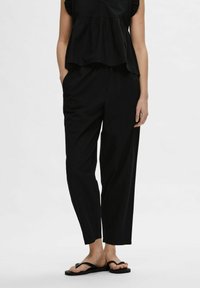 Črn peplum top z nabranimi rokavi, v kombinaciji z ohlapnimi črnimi hlačami. Model nosi črne japonke in stoji na nevtralni ozadju.