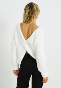 Suéter blanco y esponjoso con escote en V profundo en la espalda y detalle retorcido en la cintura, combinado con pantalones negros ajustados. Las mangas son ligeramente holgadas.