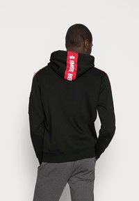 Sudadera negra con una franja vertical roja en la parte posterior con texto, y franjas laterales rojas. Tejido suave y texturizado con capucha y puños acanalados.
