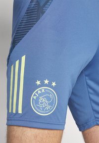 Blauwe sportshorts van gladde stof met een textuurpaneel, drie gele strepen en een Ajax Amsterdam-logo met vier sterren.