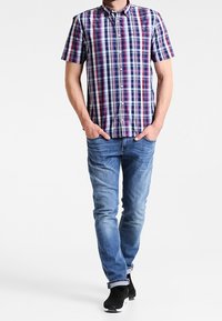 Man som går med händerna i fickorna, iförd en kortärmad flerfärgad rutig skjorta, blå jeans och svarta sneakers.