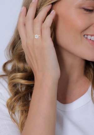 Bague en or avec une grande pierre ronde et transparente au centre, portée par une main avec des cheveux blonds ondulés et portant une chemise blanche.