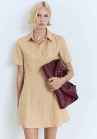 Rochie bej cu mânecă scurtă, cu guler, având o siluetă slim. Ținând o geantă de mână texturată de culoare burgundă, cu un design împletit.