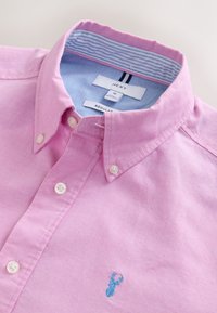 Chemise rose pâle à boutons, doublure intérieure de col bleue, boutons blancs et petit logo cerf brodé bleu sur la poitrine.