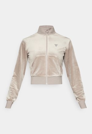 Przycięta beżowa aksamitna kurtka z wysokim kołnierzem, zamkiem błyskawicznym z przodu i elastycznymi mankietami. Posiada małe logo po lewej stronie. Miękka tekstura.