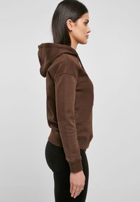Sudadera con capucha marrón, con dobladillo y puños acanalados, que presenta un cordón y bolsillos laterales, exhibida de lado sobre un fondo neutral.
