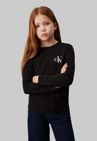 Camisola preta de manga longa com decote redondo e logótipo no peito, combinada com jeans de ganga escura. Textura do tecido suave.