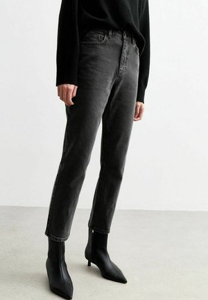 Osoba nosząca ciemnoszare jeansy z wysokim stanem i czarne, spiczaste botki stojąca na jasnoszarej podłodze.