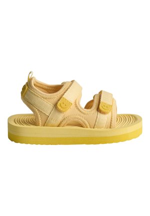 Keltainen sandaali, jossa on pehmeä, teksturoitu yläosa ja säädettävät hihnat. Pyöreä pohja, jossa on uritettu muotoilu. Hihnoissa hymyilevät kasvot koristeena.