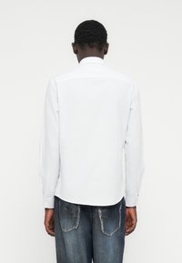 Άνδρας που στέκεται με τη πλάτη προς την κάμερα, φοράει λευκό πουκάμισο μακρυμάνικο και ξεθωριασμένο μπλε τζιν με λεπτομέρειες μπαλωμάτων στις τσέπες.