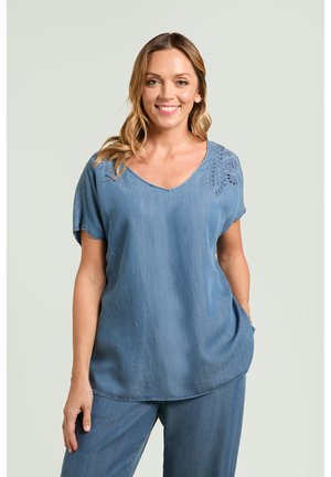 Donna sorridente con capelli mossi che indossa una maglietta blu a maniche corte con dettagli ricamati e pantaloni larghi abbinati, in piedi con una mano in tasca.
