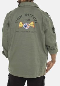 Chemise militaire verte avec un grand graphique au dos représentant un œil et des ailes, le texte "VON DUTCH 1929" et "NOUS SOMMES TOUJOURS VIVANTS", et des manches retroussées.