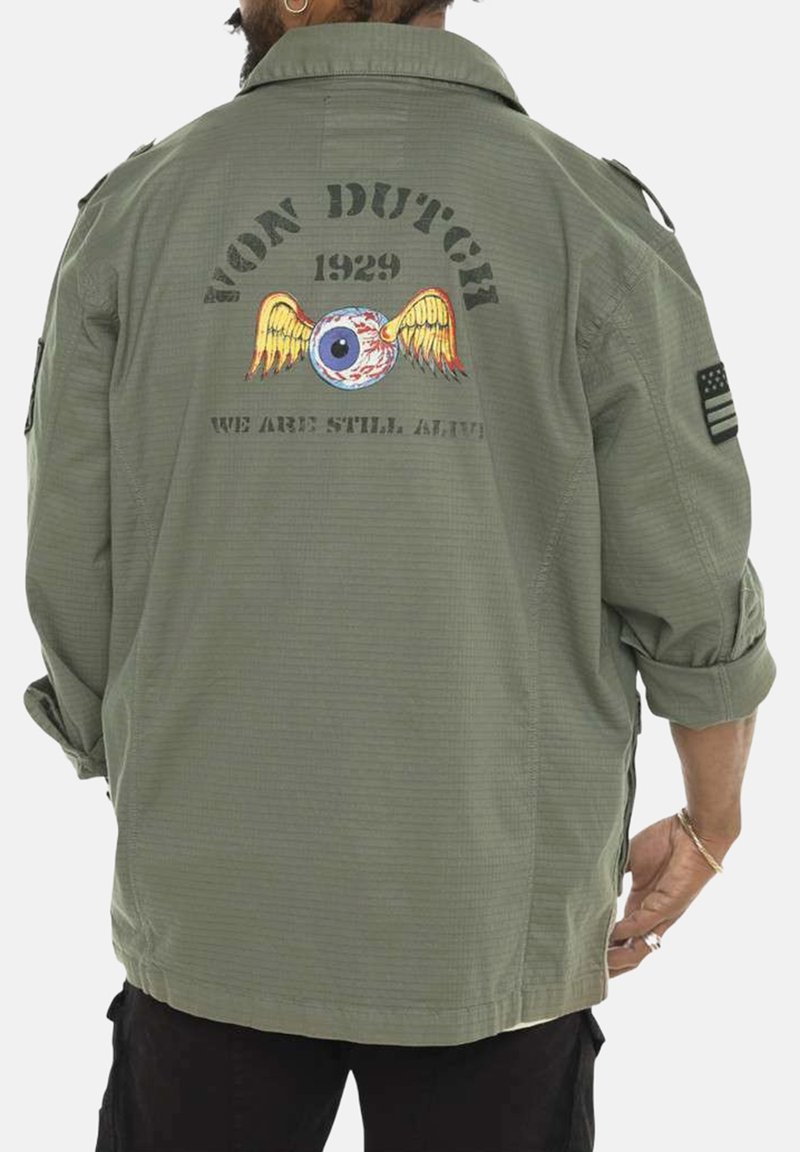 Chemise militaire verte avec un grand graphique au dos représentant un œil et des ailes, le texte "VON DUTCH 1929" et "NOUS SOMMES TOUJOURS VIVANTS", et des manches retroussées.