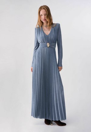 Mujer con un vestido maxi azul plisado de manga larga, con cintura ceñida y broche decorativo dorado, combinado con botas negras de tacón.