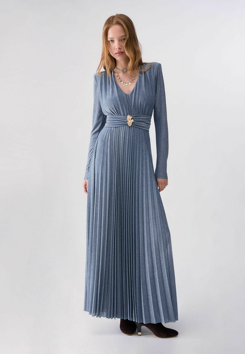 Femme portant une robe maxi bleue plissée à manches longues avec une taille cintrée et une boucle dorée décorative, associée à des bottines noires à talons.