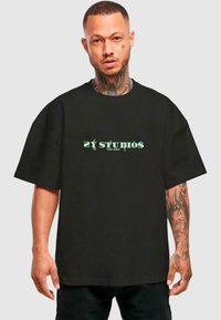 Black oversized t-shirt made of cotton with short sleeves, featuring green "2Y STUDIOS" text on the front. Model holds hands at sides.