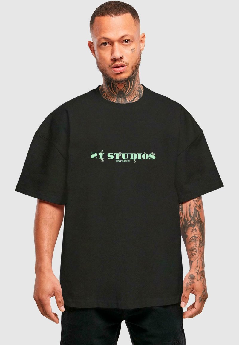 Black oversized t-shirt made of cotton with short sleeves, featuring green "2Y STUDIOS" text on the front. Model holds hands at sides.