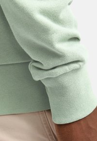 Tejido de suéter de color verde claro con detalle en puño doblado. La imagen en primer plano muestra la textura y el acabado suave, en combinación con una tela beige abajo.