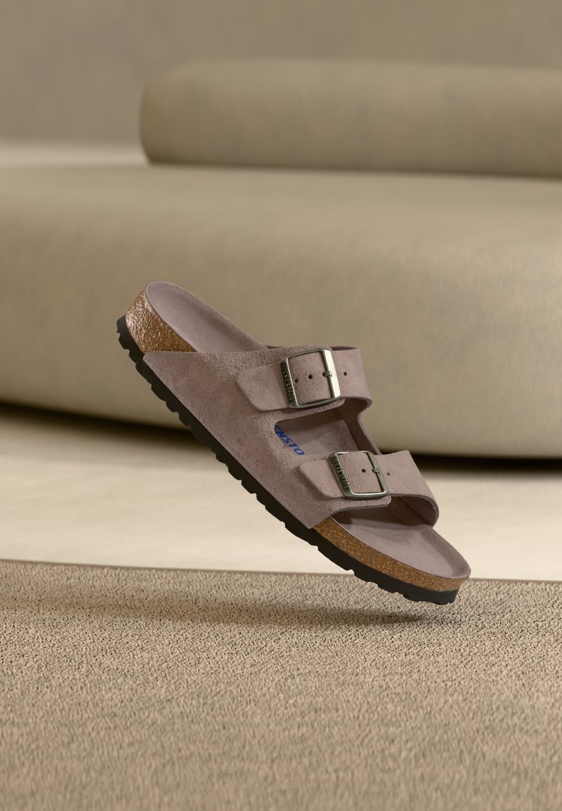 Sandalia de ante marrón claro con dos hebillas ajustables y plantilla de corcho flotando sobre una alfombra beige texturizada.