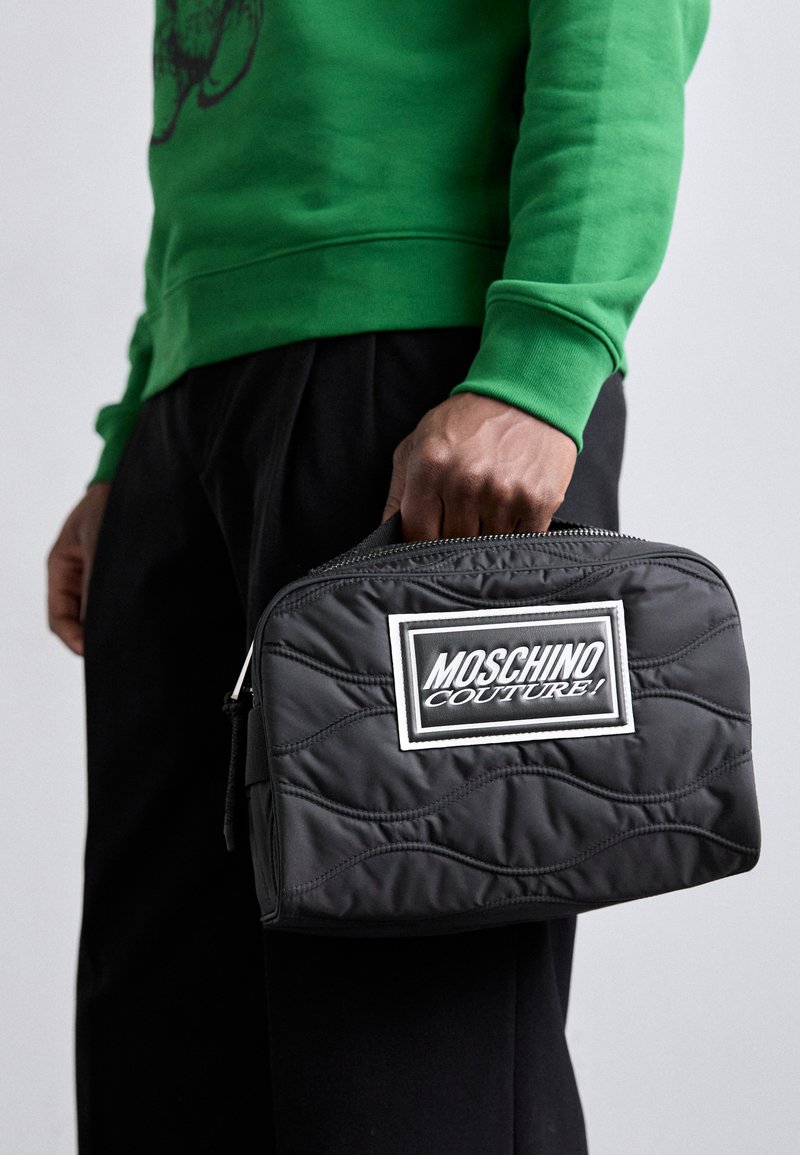 Black quilted fabric bag with a white Moschino logo patch, featuring a zip closure and a sturdy handle. Green sweatshirt worn underneath.