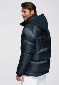 Vadderad jacka i mörk marinblå med avtagbar huva, utrustad med kviltade paneler, elastiska manschetter och en svart logotyp-patch på vänster arm.