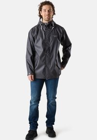 Veste grise imperméable avec capuche, boutons-pression et deux poches latérales. Portée sur un jean bleu et des chaussures noires, avec une texture lisse.