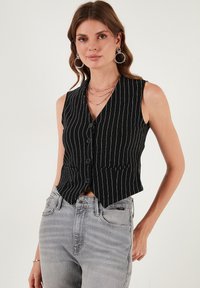 Čierno-biely pruhovaný bezrukávový sveter s výstrihom do V, zapínaním na gombíky a dvoma prednými vreckami. Nosený cez šedé džínsy s vysokým pásom.