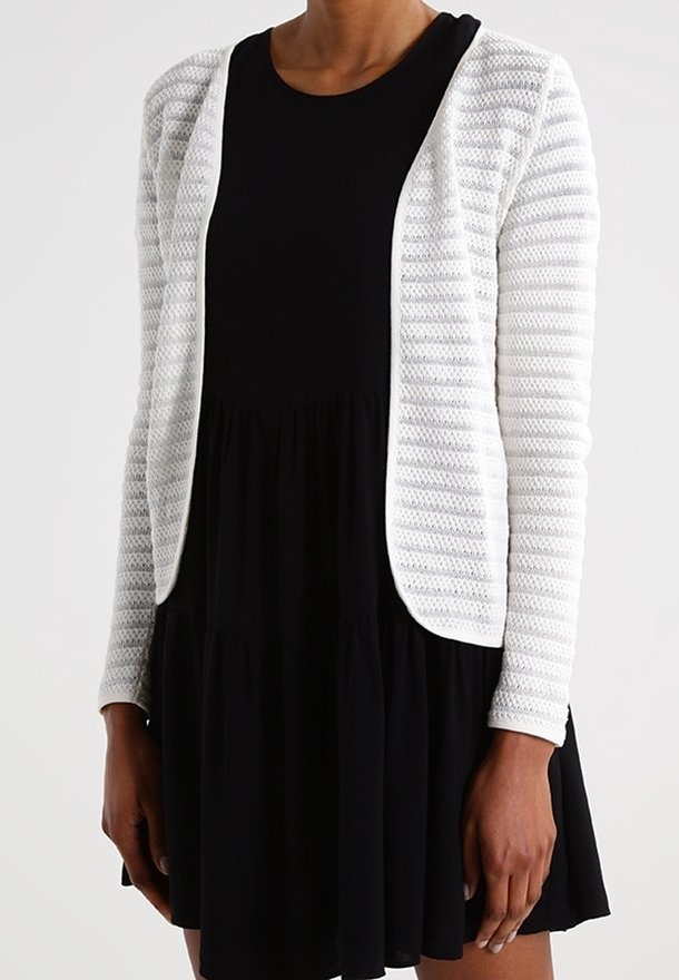 Witte gebreide cardigan met horizontale strepen, met een open voorkant. Getragen over een zwarte jurk met een uitlopende rok.