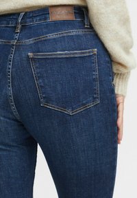 Jeans de mezclilla azul oscuro con una textura suave, que presentan dos bolsillos traseros, costuras amarillas contrastantes y una etiqueta de cuero marrón en la cintura.