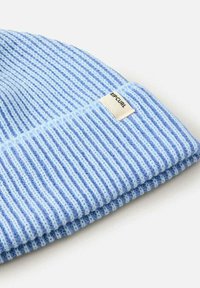 Gorro de punto azul claro con franjas alternas en azul oscuro y azul claro. Cuenta con una etiqueta rectangular blanca con la inscripción "RIP CURL". Textura suave.
